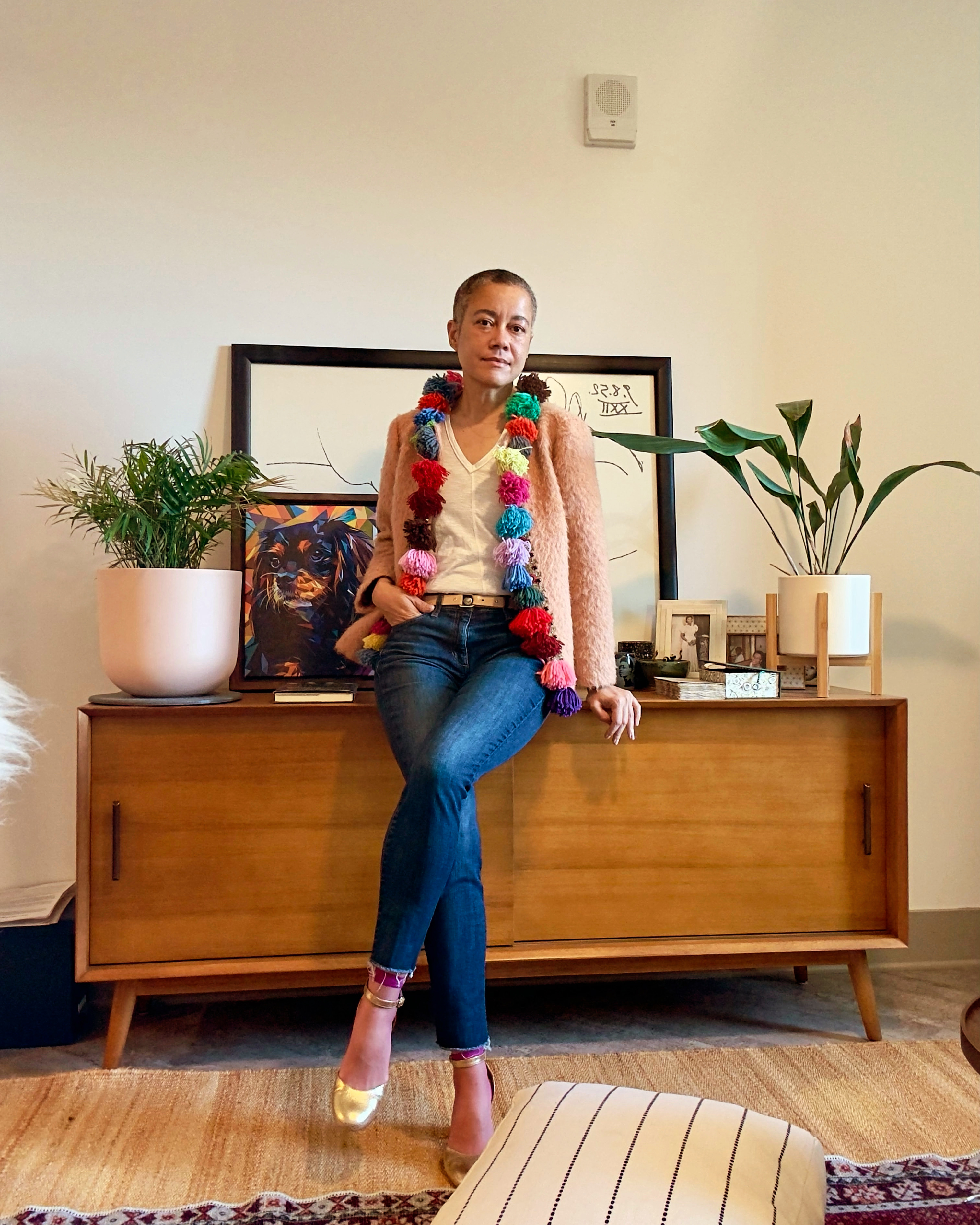 A casually dressed person sitting on a wooden sideboard, wearing a colorful pom-pom trimmed jacket, with a plant and framed artwork in the background.